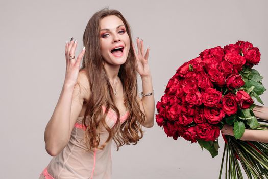 Studio portrait of stunning young brunette woman with make up wearing sexy night dress smelling a big bunch of red roses with closed eyes. Enjoying the smell of roses.