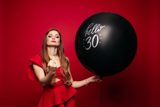 Studio portrait of attractive self-confident brunette woman in stylish red dress with black air balloon says hello 30 showing fuck at camera. Isolate on red background.
