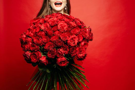 Selective focus of beautiful bouquet of red roses in hands of happy woman. Young lady laughing and keeping wonderful flowers on red isolated background. Concept of valentines day and present.