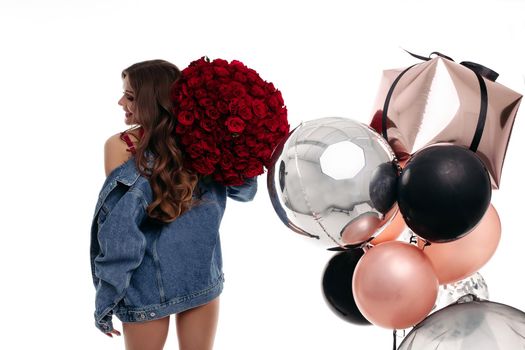 Woman's back in blue jeans jacket holding big bouquet of roses on her shoulder. Brunette girl with long hair getting presents on birthday. Stylish lady standing near gold, silver and black balloons.