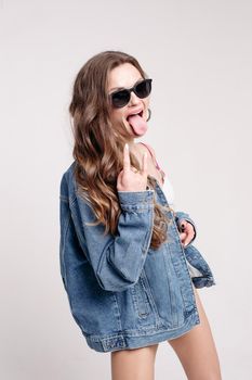 Studio portrait of positive funny brunette teen or adult girl in black sunglasses in trendy denim jacket showing rock and roll gesture with her hand. standing over white background.