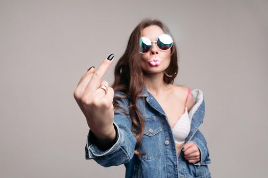 Offensive gesture showing at camera. Stylish young brunette in mirrored sunglasses showing middle finger with black nail. She is wearing denim jacket over a bra. Isolate on grey. Studio shot.