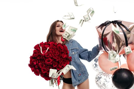 Funny woman in jeans jacket laughing loudly while money falling around. Attractive model keeping bouquet of red roses and pink balloons and posing on isolated background. Concept of girt and surprise.