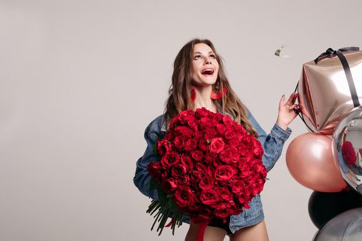Funny woman in jeans jacket laughing loudly while money falling around. Attractive model keeping bouquet of red roses and pink balloons and posing on isolated background. Concept of girt and surprise.
