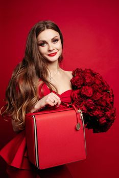 Fashionable studio portrait of stylish young woman with long brown hair in expensive red suit with leather red bag and bunch of red roses. She is smiling at camera. Red lips. Red background.