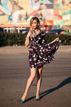 Full length portrait of stunning blonde woman in colored black, white and red dress and green heels posing with skirt in hand and eyes closed in bright sunshine in the street. Colored background.