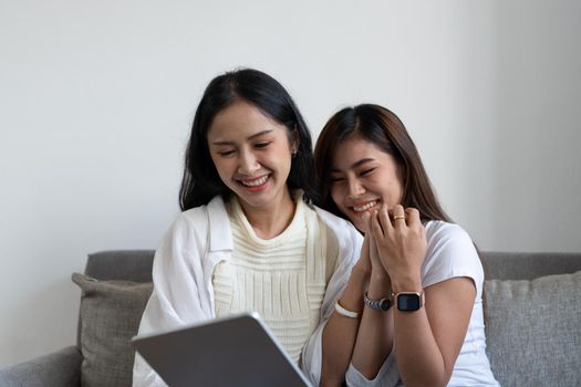 Happy young asian lesbian couple hugging having fun using digital tablet relaxing on couch at home. Two smiling women friends holding computer looking at screen enjoying surfing online watching videos.