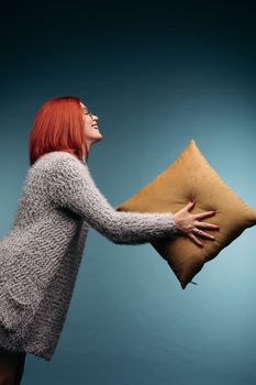 Portrait of red haired woman in gray sweater holding by hands and giving at side yellow pillow, posing on blue background. Pretty girl caring about her dreams and comfort sleeping. Concept of decor.