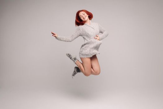 Happy woman jumping and holding hand on belly, with other showing blanket space. Charming and adorable model with ginger hair posing in studio with grey background. Girl wearing in grey sweater.