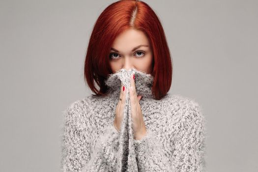 Studio portrait of young red haired unrecognizable woman wearing warm gray seater hiding face by hands. Incognito girl having reaction of protection or saving secret. Gray studio background.