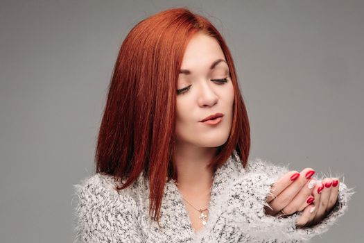 Beautiful young girl with red hair and red nails holding her hands together and blowing on white lightening ball. Pretty woman in grey sweater catching star and making wishes when snowing.
