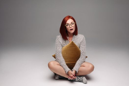 Red haired girl in grey sweater and warm socks sitting on floor in Turkish style and sadly looking at camera. Beautiful girl in glasses with cross on her neck holding pillow between hands and legs.