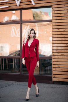 Pretty elegance stylish lady in red suit and black shoes posing at camera. Young gorgeous girl standing with crossing legs on street near caffee. Sexy blond woman walking in sunny day.