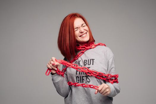 Studio portrait of a girl entangled in a chain. A red-haired woman with brown hair tries to get rid of the chain. The concept of a life-like situation. Isolated on a gray background.