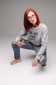 Swag and confident red haired girl in jeans and sweatshirt posing at gray studio, looking at camera and gesturing by hands. Young stylish woman wearing in bag girl look. Concept of fashion.