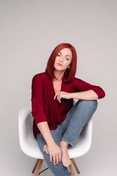 Beautiful young woman wearing in marsala blouse and jeans sitting on white chair. Model with straight ginger hair looking at camera with serious face. Girl posing in studio with grey background.
