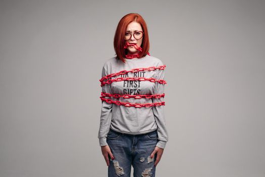 Studio portrait of a girl entangled in a chain. A red-haired woman with brown hair tries to get rid of the chain. The concept of a life-like situation. Isolated on a gray background.
