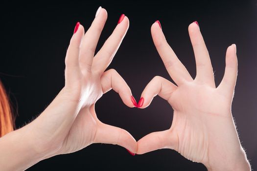 Crop of two hadns together showing heart by fingers and gesturing symbol of amour. Isolated love sign making by hands with red manicured nails on black studio background.
