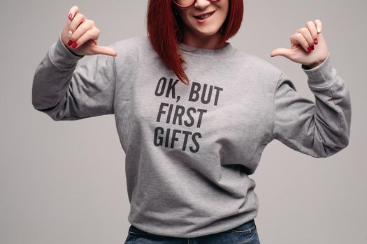Crop of fashionable red haired girl wearing gray sweatshirt with description about for first gifts. Stylish woman posing in studio pointing by fingers, showing her look, waiting for presents.