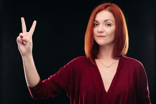 Attractive red haired girl in bordo blouse thinking about something and holding hand with two finger up. Young beautiful woman on black background seriously looking at camera. Pretty lady having new idea.