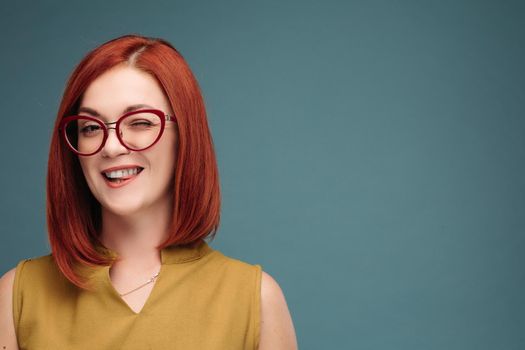 Studio portrait of a happy girl with brown hair. A red-haired woman in stylish glasses smiles looking at the camera. Close-up. Isolated on a blue background