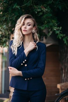 Beautiful blond girl with long neck and big flower shaped earings looking seriously at camera. Young sexy pretty woman wearing dark blue suit with big gold buttons and holding one hand near breast.