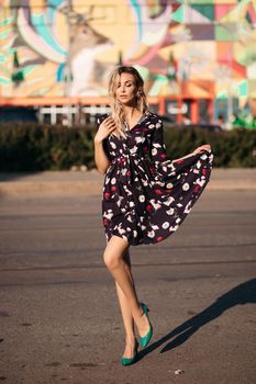 Full length portrait of stunning blonde woman in colored black, white and red dress and green heels posing with skirt in hand and eyes closed in bright sunshine in the street. Colored background.
