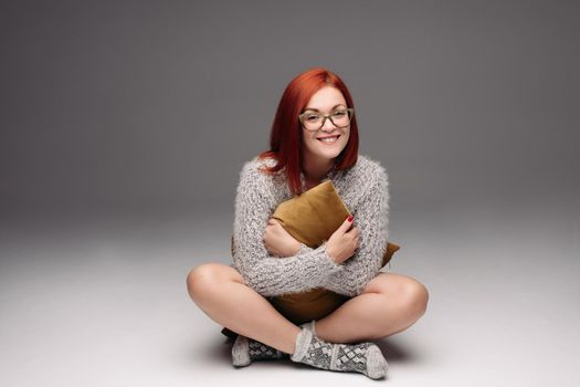 Red haired girl in grey sweater and warm socks sitting on floor in Turkish style and sadly looking at camera. Beautiful girl in glasses with cross on her neck holding pillow between hands and legs.