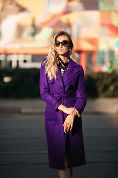 Young stylish woman in sunglasses posing at camera in sunny day. Blond girl in black blouse and violet coat holding her hands pockets. Elegant pretty lady in fashionable clothes standing outside.