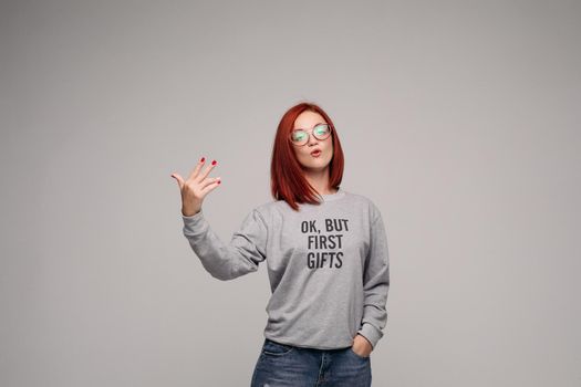 Swag and confident red haired girl in jeans and sweatshirt posing at gray studio, looking at camera and gesturing by hands. Young stylish woman wearing in bag girl look. Concept of fashion.