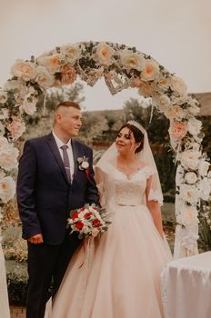 Bride and groom in a wedding dress and a long veil at a wedding ceremony on the background of a flower arch laughing kissing rejoicing. Slavic Ukrainian Russian traditions
