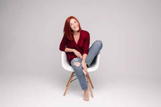Beautiful young woman wearing in marsala blouse and jeans sitting on white chair. Model with straight ginger hair looking at camera with serious face. Girl posing in studio with grey background.