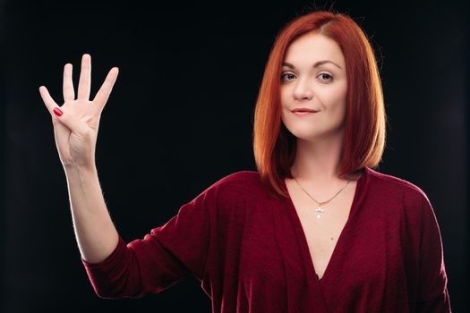 Attractive red haired girl in bordo blouse thinking about something and holding hand with four finger up. Young beautiful woman on black background seriously looking at camera. Pretty lady having new idea.