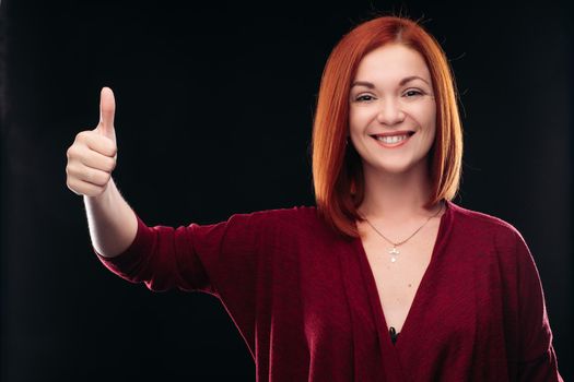 Attractive red haired girl in bordo blouse thinking about something and holding hand with finger up. Young beautiful woman on black background seriously looking at camera. Pretty lady having new idea.