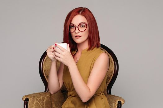 Studio portrait of a beautiful luxury girl with chestnut hair. Red-haired girl is seated on a chair in a sexy suit and heels. Isolated on a gray background.