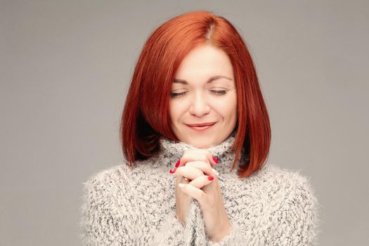 Adorable and beautiful young woman with smooth skin and straight ginger hair posing. Beautiful model looking at camera and smiling. Girl wearing in grey sweater and putting hands together.