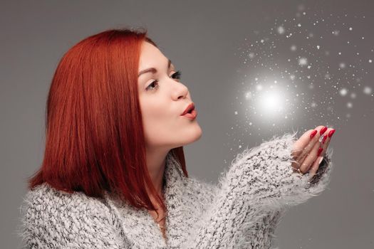 Beautiful young girl with red hair and red nails holding her hands together and blowing on white lightening ball. Pretty woman in grey sweater catching star and making wishes when snowing.