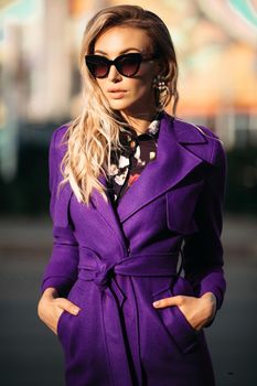 Young beautiful stylish woman in sunglasses posing at camera in sunny day. Blond girl in black blouse and violet coat standing and holding her hands in pockets. Concept of style and fashion.