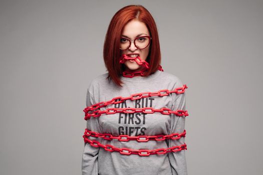 Studio portrait of a girl entangled in a chain. A red-haired woman with brown hair tries to get rid of the chain. The concept of a life-like situation. Isolated on a gray background.