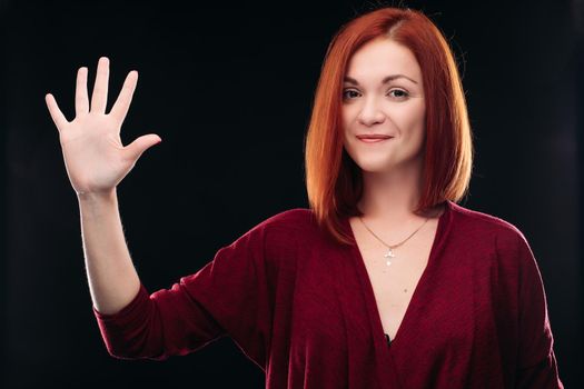 Attractive red haired girl in bordo blouse thinking about something and holding hand with five finger up. Young beautiful woman on black background seriously looking at camera. Pretty lady having new idea.