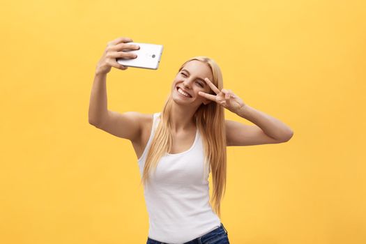 Self portrait of charming cheerful girl shooting selfie on front camera gesturing v-sign peace symbol with fingers isolated on yellow background.