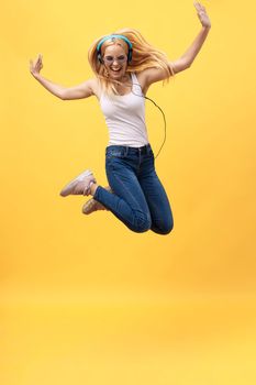 Full-length portrait of carefree woman in jean jumping while listening music. Indoor photo of adorable caucasian female model in white t-shirt fooling around in studio with yellow background