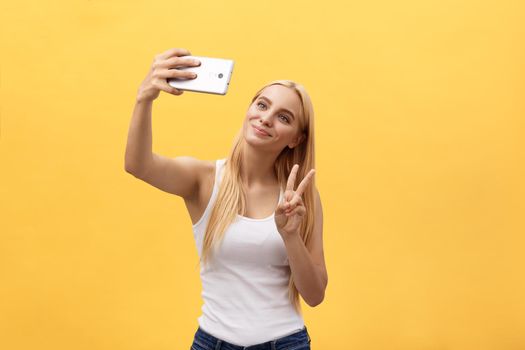 Self portrait of charming cheerful girl shooting selfie on front camera gesturing v-sign peace symbol with fingers isolated on yellow background.