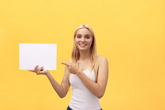 Young woman over yellow background holding blank paper sheet with surprise face pointing finger.