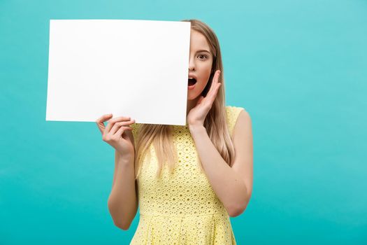 Portrait of amazed young blond woman holding blank sign with copy space on blue studio background. Showing shocked surprise face