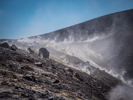 Hilly uneven slopes with rough boulders and smoke, on volcanic island Vulcano, located in Southern Italy in sunny day with blue sky