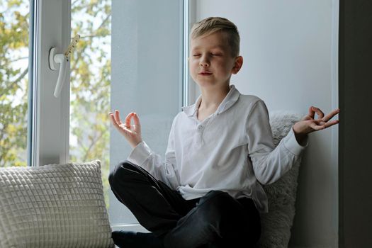 Cute school boy in white shirt reading sitting on a windowsill and meditatig