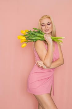 Adult blonde woman in pink dress posing over rosy background. Spring-summer clothing. She is holding bouquet of yellow tulips in her hands. Presents, surprise