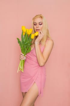 Adult blonde woman in pink dress posing over rosy background. Spring-summer clothing. She is holding bouquet of yellow tulips in her hands. Presents, surprise