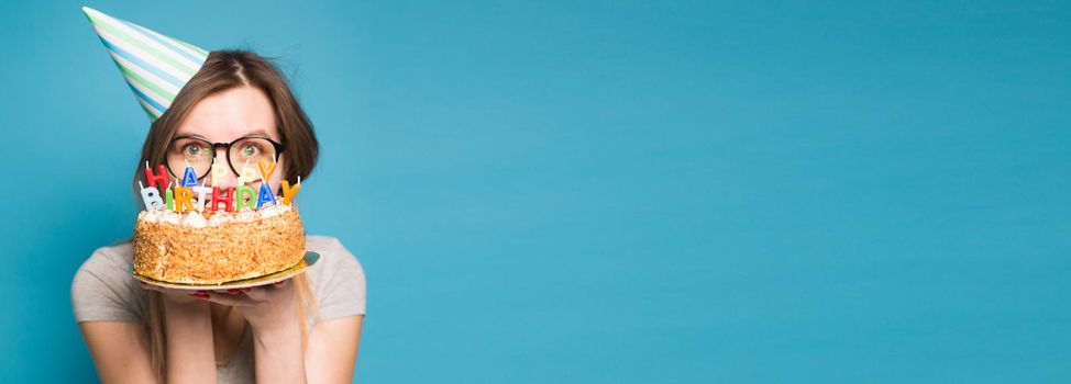 Close up funny blurred positive girl in glasses and greeting paper hat holding a happy birthday cake in her hands standing on a blue background. Concept of congratulations and anniversaries. Copyspace